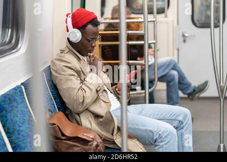 Afro-American passenger man in red hat, trench coat siting in subway train, using mobile phone, listens to music with wireless headphones in public tr Stock Photo