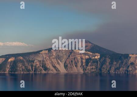 Thick smoke from a wildfire billows up into the sky behind Mt. Scott and the eastern rim of Crater Lake as afternoon sunlight casts a soft glow. Stock Photo