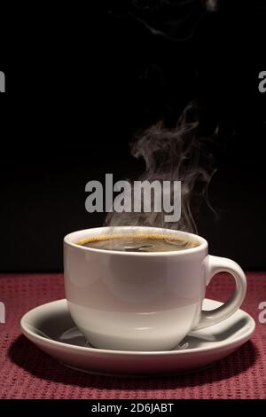 Coffee steaming in a cup with saucer isolated on a black background on a red surface Stock Photo