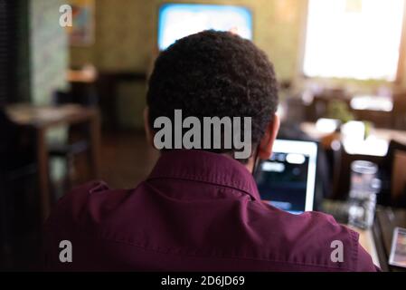 Young black male  developer sitting in pizza restaurant, drinking coffee or  tea and working with laptop. Stock Photo