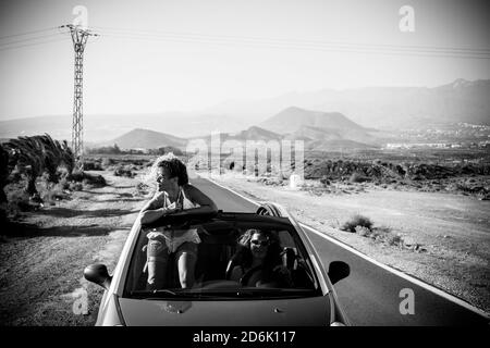 Black and white conceptual travel and adventure with friends concept with couple of young middle age women traveling on a convertible car in a long ro