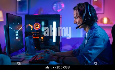 Excited Gamer Playing First-Person Shooter Online Video Game on His Personal Computer. Room and PC have Colorful Neon Led Lights. Young Man has Long Stock Photo