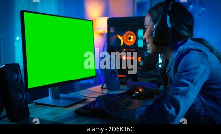 Pretty and Excited Black Blogger Girl in Headphones is Using PC with Green Screen Mock Up. Room and PC have Colorful Neon Led Lights. Cozy Evening at Stock Photo