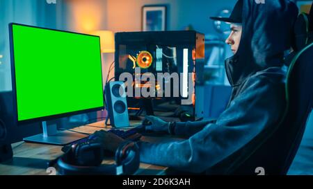 Professional Stream Using His Cool Personal Computer with Green Screen Mock-up Template. Young Man is Wearing a Cap and Hood. Room and PC have Stock Photo