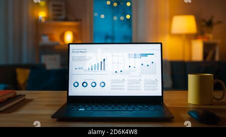 Laptop Computer Showing Statistical Infographics Stands on a Desk in the Living Room. In the Background Cozy Living Room with Warm Lights on. Stock Photo