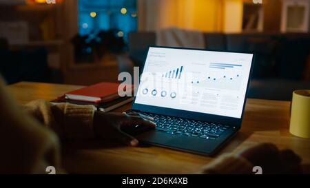 Laptop Computer Showing Statistical Infographics Stands on a Desk in the Living Room. In the Background Cozy Living Room with Warm Lights on. Stock Photo