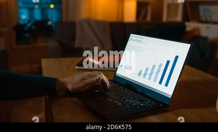 Over the Shoulder Shot: Young Man Sitting at His Desk Works on a Laptop that Shows Statistics and Infographics. Late at Night in His Living Room Man Stock Photo