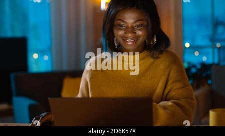 Portrait of Beautiful Smiling Black Girl Working on a Laptop while Sitting at Her Desk at Home. In the Evening Creative Woman Works on a Computer In Stock Photo