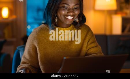 Portrait of Beautiful Smiling Black Girl Working on a Laptop while Sitting at Her Desk at Home. In the Evening Creative Woman Works on a Computer In Stock Photo