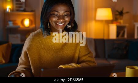 Portrait of Beautiful Smiling Black Girl Working on a Laptop while Sitting at Her Desk at Home. In the Evening Creative Woman Works on a Computer In Stock Photo