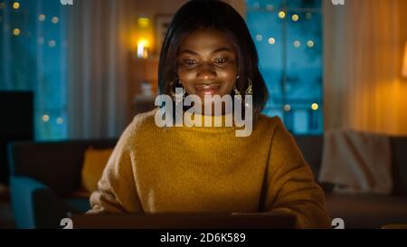 Portrait of Beautiful Black Girl Working on a Laptop while Sitting at Her Desk at Home. In the Evening Creative Woman Works on a Computer In Her Cozy Stock Photo