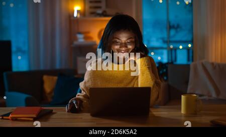 Portrait of Beautiful Black Girl Uses Computer while Sitting at Her Desk at Home, She's Wearing Warm Sweater. In the Evening Creative Woman Works on a Stock Photo