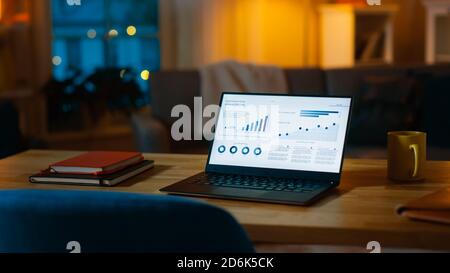 Laptop Computer Showing Statistical Infographics Stands on a Desk in the Living Room. In the Background Cozy Living Room with Warm Lights on. Stock Photo