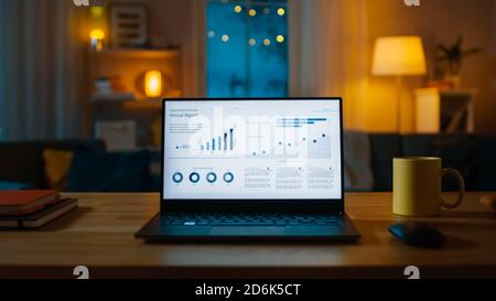 Laptop Computer Showing Statistical Infographics Stands on a Desk in the Living Room. In the Background Cozy Living Room with Warm Lights on. Stock Photo