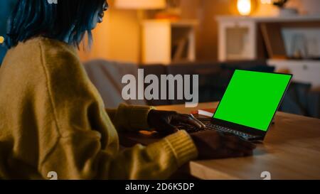 Beautiful Black Woman Sitting at Her Desk Works on a Laptop with Green Mock-up Screen. Late at Night in Her Living Room Girl Uses Notebook Computer. Stock Photo