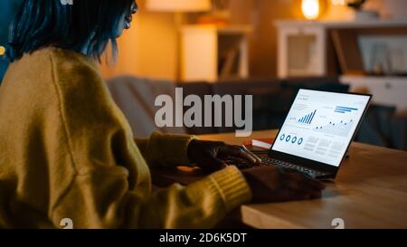 Laptop Computer Showing Statistical Infographics Stands on a Desk in the Living Room. In the Background Cozy Living Room with Warm Lights on. Stock Photo