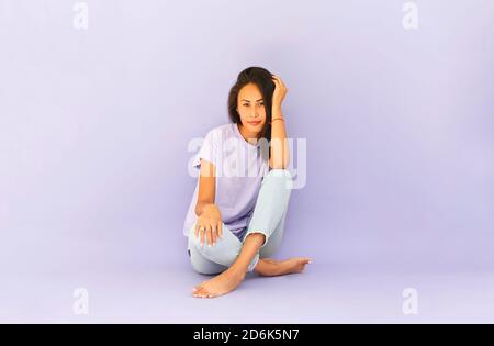 Full body of positive barefoot millennial Asian female in casual shirt and jeans sitting on floor in light violet studio Stock Photo