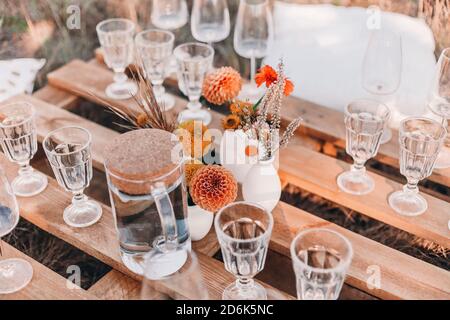 Set of elegant glassware placed on plank wooden table with floral arrangement on grassy meadow during festive autumn picnic Stock Photo