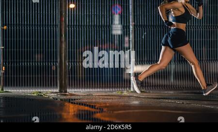 Beautiful Energetic Fitness Girl is Sprinting in a Fenced Outdoor Basketball Court. She's Running at Night After Rain in a Residential Neighborhood Stock Photo