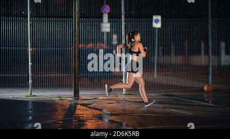 Beautiful Energetic Fitness Girl is Sprinting in a Fenced Outdoor Basketball Court. She's Running at Night After Rain in a Residential Neighborhood Stock Photo