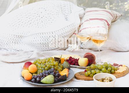 Pair of glasses with wine placed near tables with various fresh ripe fruits on blanket with cushions during romantic picnic in garden Stock Photo