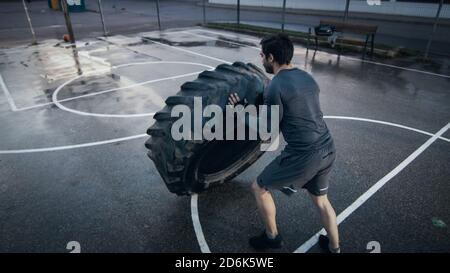 Strong Muscular Fit Young Man in Sport Outfit and Gloves is Doing Exercises in a Fenced Outdoor Basketball Court. He's Flipping a Big Heavy Tire in an Stock Photo