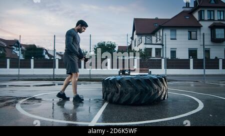 Strong Muscular Fit Young Man in Sport Outfit and Gloves is Doing Exercises in a Fenced Outdoor Basketball Court. He's Looking at Big Heavy Tire in an Stock Photo