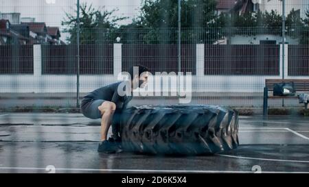 Strong Muscular Fit Young Man in Sport Outfit and Gloves is Doing Exercises in a Fenced Outdoor Basketball Court. He's Flipping a Big Heavy Tire in an Stock Photo