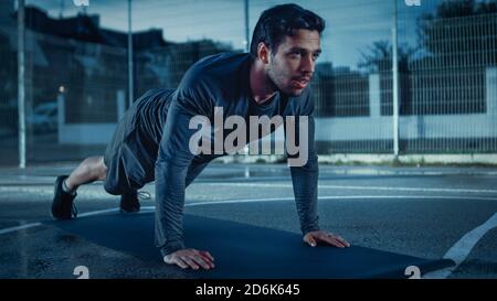 Strong Muscular Fit Young Man in Sport Outfit Doing Push Up Exercises. He is Doing a Workout in a Fenced Outdoor Basketball Court. Evening Shot After Stock Photo