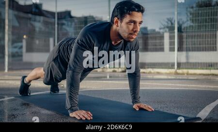 Strong Muscular Fit Young Man in Sport Outfit Doing Push Up Exercises. He is Doing a Workout in a Fenced Outdoor Basketball Court. Evening After Rain Stock Photo