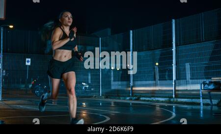 Beautiful Energetic Fitness Girl is Sprinting in a Fenced Outdoor Basketball Court. She's Running at Night After Rain in a Residential Neighborhood Stock Photo