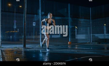 Beautiful Energetic Fitness Girl is Sprinting in a Fenced Outdoor Basketball Court. She's Running at Night After Rain in a Residential Neighborhood Stock Photo