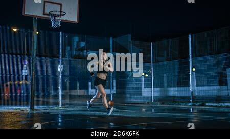 Beautiful Energetic Fitness Girl is Sprinting in a Fenced Outdoor Basketball Court. She's Running at Night After Rain in a Residential Neighborhood Stock Photo