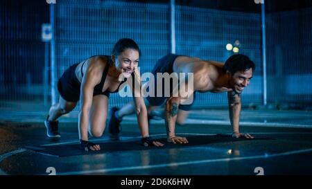 Smiling Happy Athletic Fitness Couple Doing Mountain Climber Exercises. Workout is Done in a Fenced Outdoor Basketball Court. Night After Rain in a Stock Photo