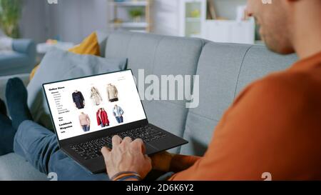 Handsome Young Man Sits at Home Uses Laptop and Browses Through Online Retailer Clothes Selling Store. Man Does Online Shopping for Clothing Items as Stock Photo