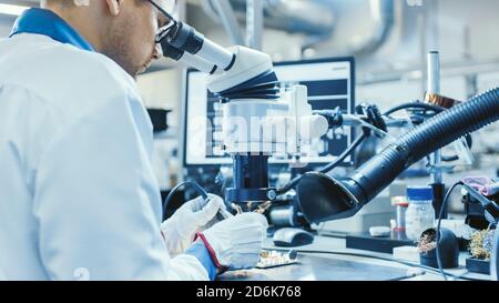 Electronics Factory Worker in White Work Coat is Soldering a Printed Circuit Board Through a Digital Microscope. High Tech Factory Facility. Stock Photo