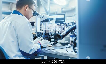 Electronics Factory Worker in White Work Coat is Soldering a Printed Circuit Board Through a Digital Microscope. High Tech Factory Facility. Stock Photo