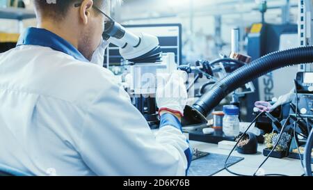 Electronics Factory Worker in White Work Coat is Soldering a Printed Circuit Board Through a Digital Microscope. High Tech Factory Facility. Stock Photo