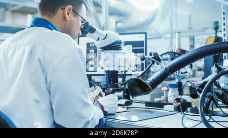 Electronics Factory Worker in White Work Coat is Soldering a Printed Circuit Board Through a Digital Microscope. High Tech Factory Facility. Stock Photo