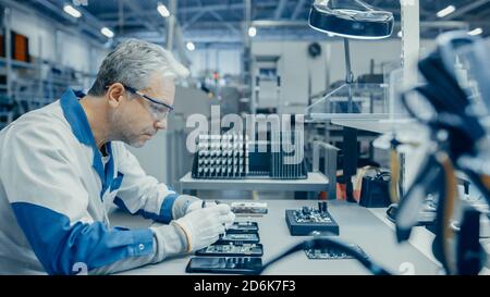 Senior Man in Blue - White Work Coat is Using Plier to Assemble Printed Circuit Board for Smartphone. Electronics Factory Workers in a High Tech Stock Photo