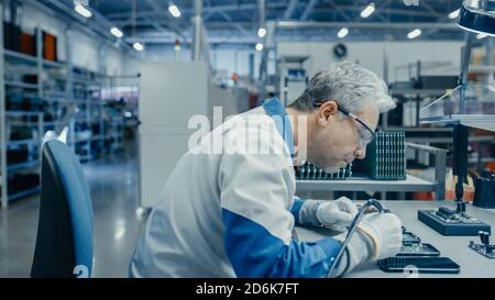 Senior Man in Blue - White Work Coat is Using Plier to Assemble Printed Circuit Board for Smartphone. Electronics Factory Workers in a High Tech Stock Photo