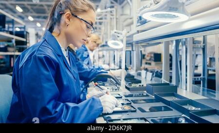 Woman Electronics Factory Worker in Blue Work Coat and Protective Glasses is Assembling Smartphones with Screwdriver. High Tech Factory Facility with Stock Photo