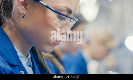 Close-up Shot of a Female Electronics Factory Worker in Blue Work Coat and Protective Glasses is Looking down and Working. High Tech Factory Facility Stock Photo
