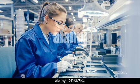 Woman Electronics Factory Worker in Blue Work Coat and Protective Glasses is Assembling Smartphones with Screwdriver. High Tech Factory Facility with Stock Photo