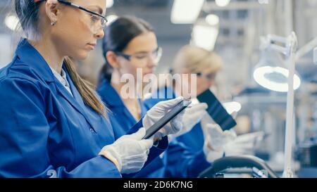 Woman Electronics Factory Worker in Blue Work Coat and Protective Glasses Takes a Smartphone Screen and Performs a Quality Check. High Tech Factory Stock Photo