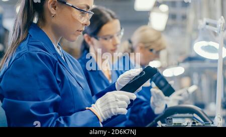 Woman Electronics Factory Worker in Blue Work Coat and Protective Glasses Takes a Smartphone Screen and Performs a Quality Check. High Tech Factory Stock Photo
