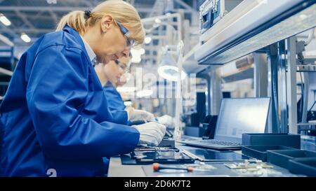Female Electronics Factory Worker in Blue Work Coat and Protective Glasses is Assembling Smartphones with Screwdriver. High Tech Factory Facility with Stock Photo