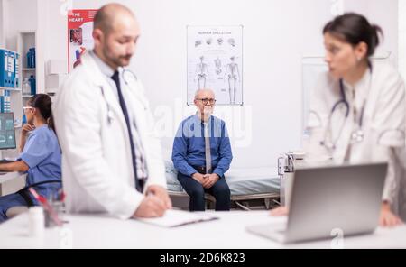 Two specialists doctors discussing senior patient diagnosis in hospital office. Scared mature man waiting medic expertise for his illness. Nurse working on laptop. Stock Photo