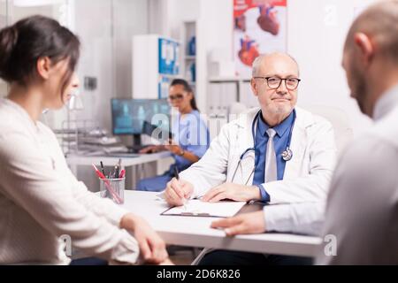 Senior medical practitioner taking notes on clipboard during consultation of married couple in hospital office. Doctor looking at male patient while wearing white coat and stethoscope. Stock Photo