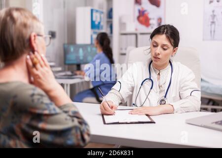 Doctor in hospital office writing treatment for senior woman during examination. Nurse wearing blue uniform working on computer. Stock Photo